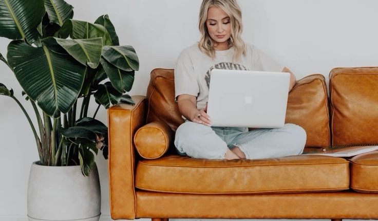 remote worker sits on a leather sofa next to a large plant