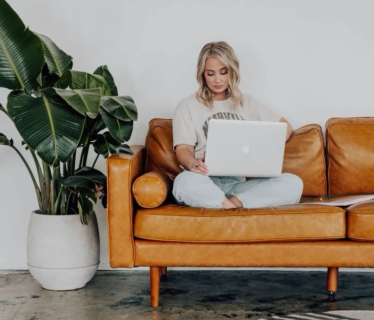 remote worker sits on a leather sofa next to a large plant