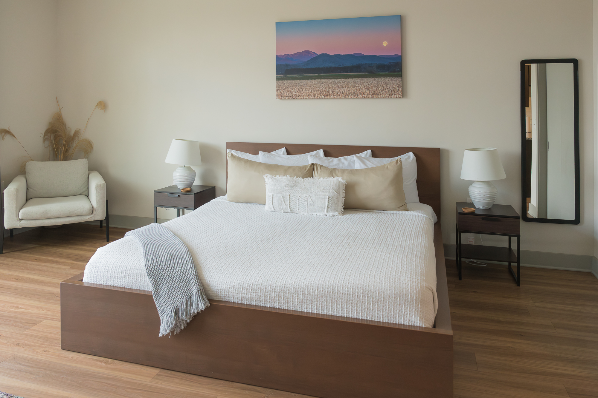 A neatly made bed with white and beige linens, flanked by two nightstands with lamps, in a room with a chair and a mirror.