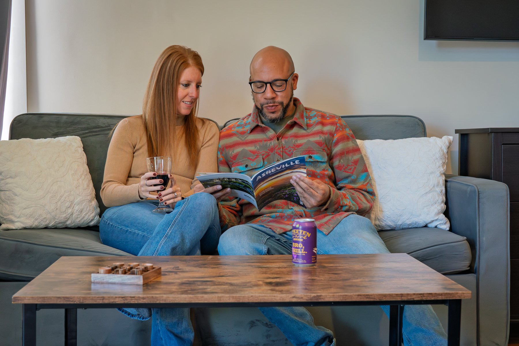 A couple sitting on a couch in a hotel lobby, reading a book and holding a glass of wine.